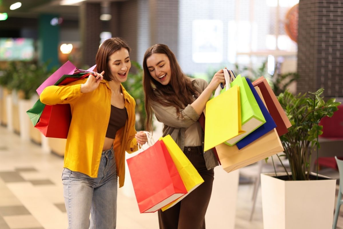 hai cô gái đang cười và cầm nhiều túi mua sắm đầy màu sắc trong trung tâm thương mại