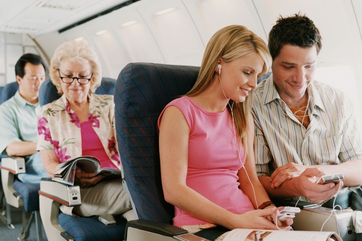 Two young people sitting on the plane looking at their phones, other passengers reading magazines
